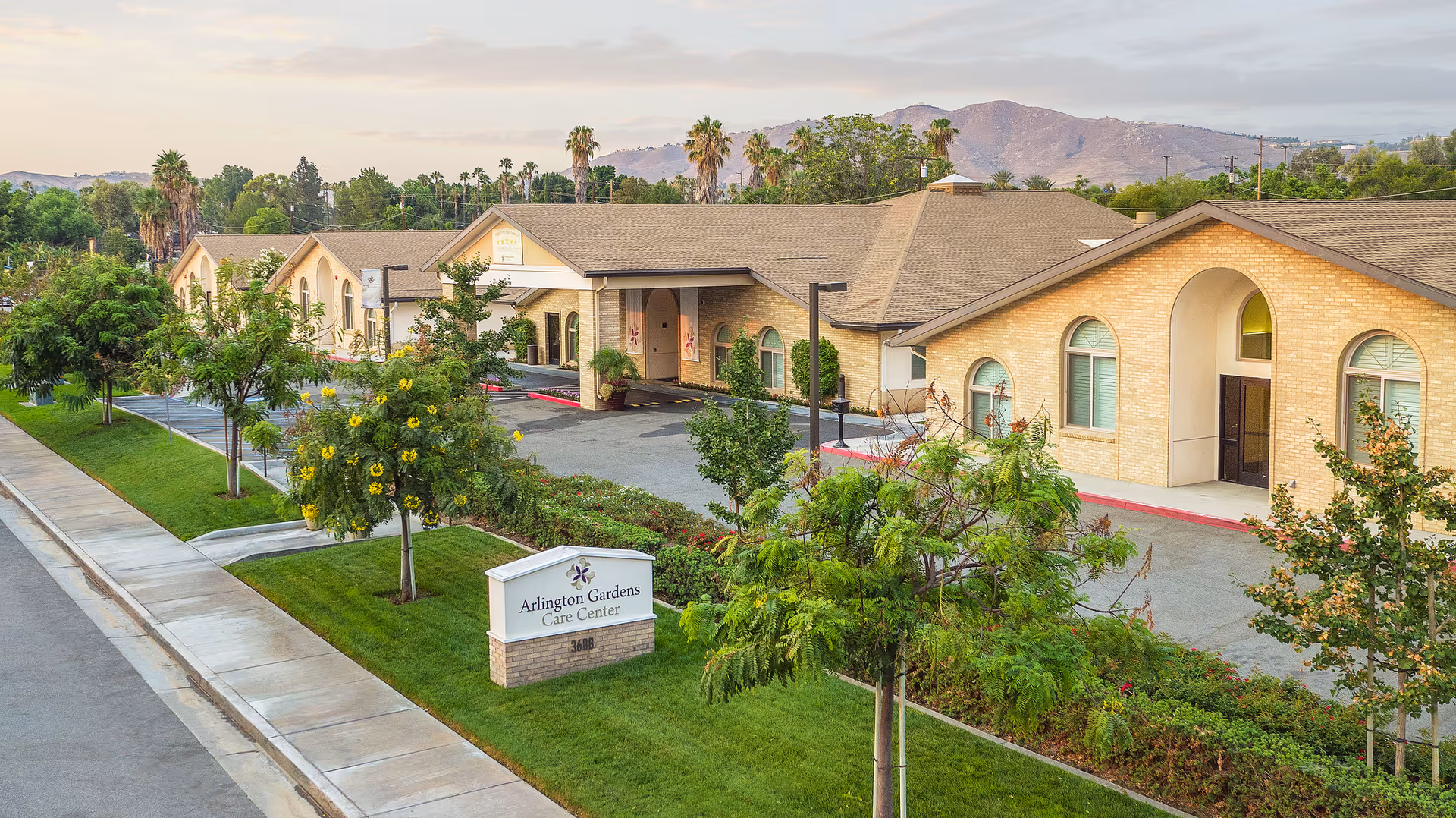 Exterior view of Arlington Gardens Care Center with beige brick building, sign, trees, and distant mountains.