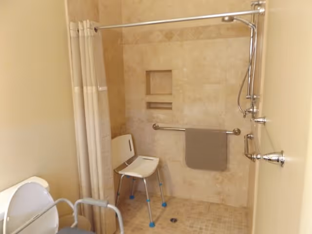 Accessible shower area with chair, grab bars, curtain, and beige tiled walls.