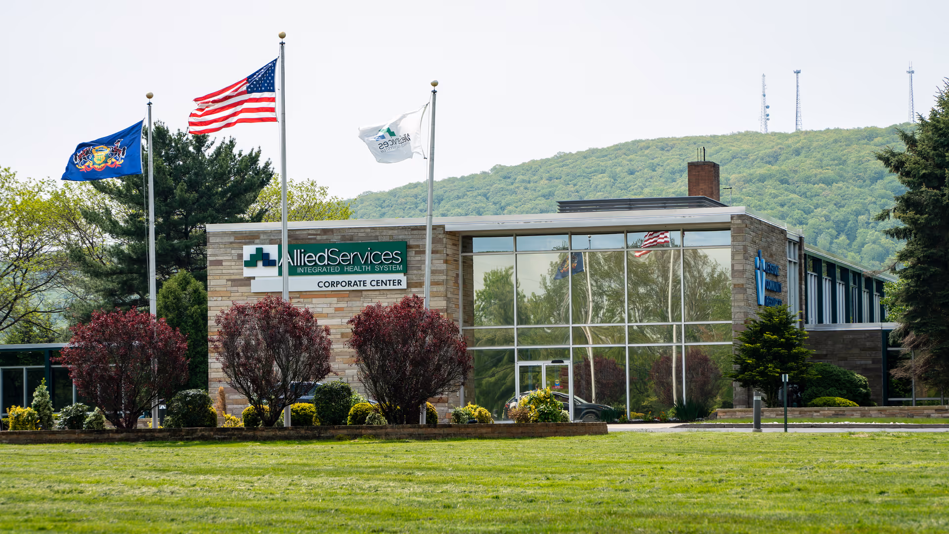 Facade of Allied Services building with flags, glass windows, and a landscaped front lawn.
