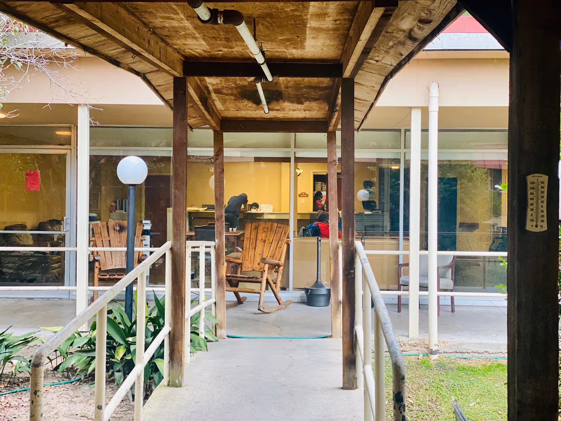 Covered outdoor walkway with wooden posts leading to a glass-fronted common area where chairs and a person at a desk are visible.