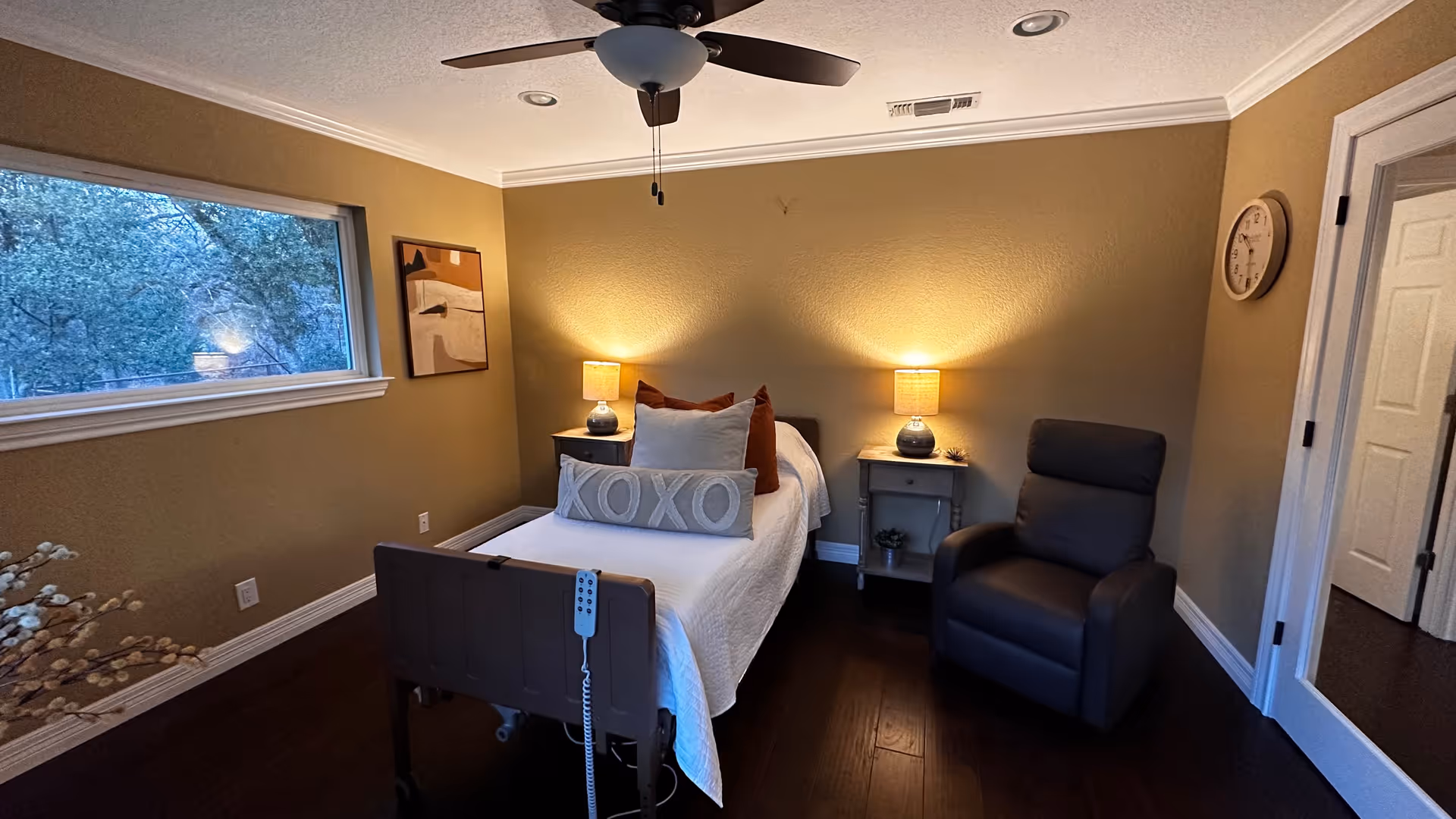 Cozy bedroom in a senior living facility with a bed, recliner, and warm lamps.