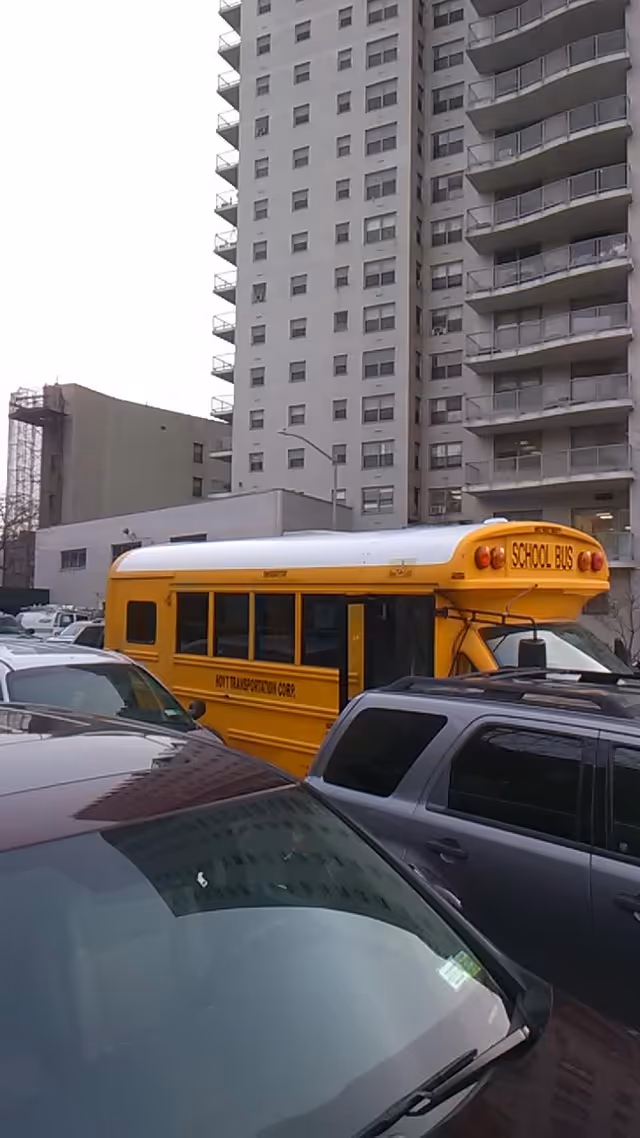 Yellow school bus parked beside a tall high-rise building with balconies.