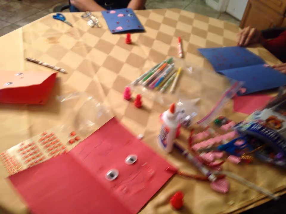A table covered with a checkered tablecloth displaying various craft supplies including glue, markers, scissors, googly eyes, and colored paper with faces drawn on them. Several hands are visible working on the crafts.