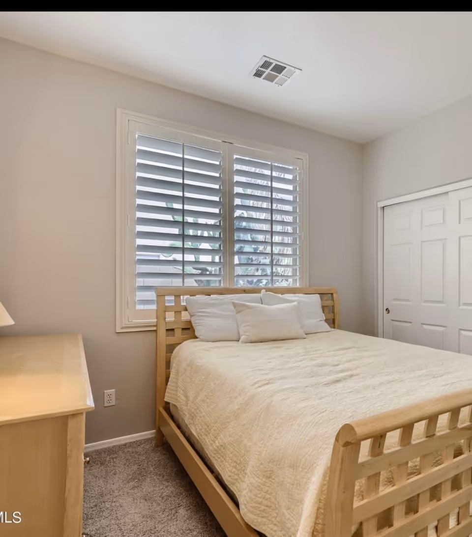 Light wood bed with white quilted bedding sits in a bright bedroom with window shutters