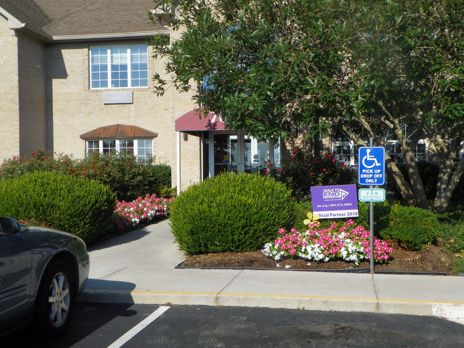 Exterior view of Liberty Ridge Senior Living showing the entrance with a maroon awning, surrounded by green bushes and colorful flowers. A car is parked in the foreground near a sidewalk leading to the entrance. There are signs indicating a handicapped pick-up and drop-off area and a purple sign for the Walk to End Alzheimer's event.