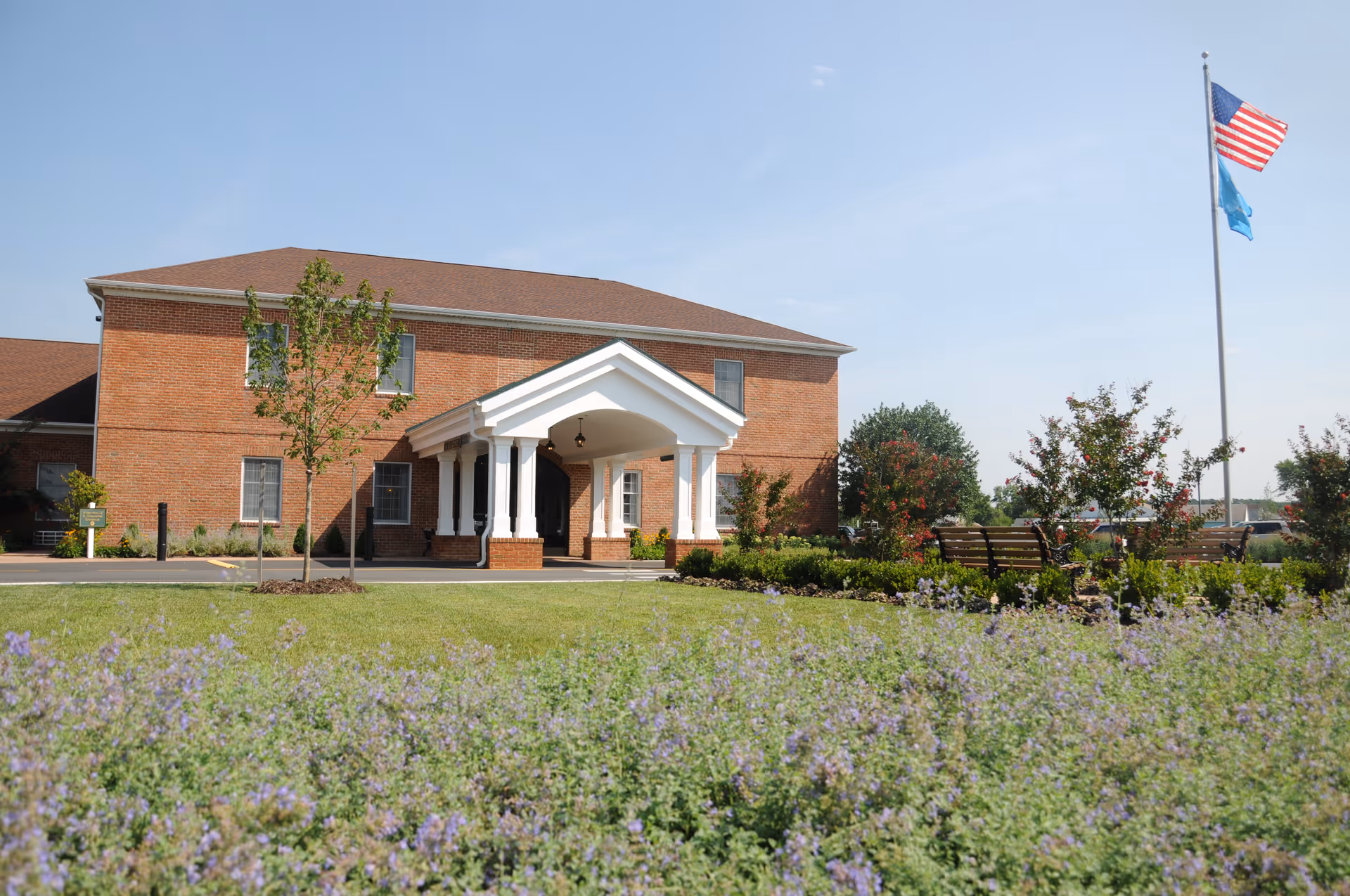 Front exterior of a two-story brick rehabilitation center with a covered entrance, landscaped lawns and an American flag on a flagpole.