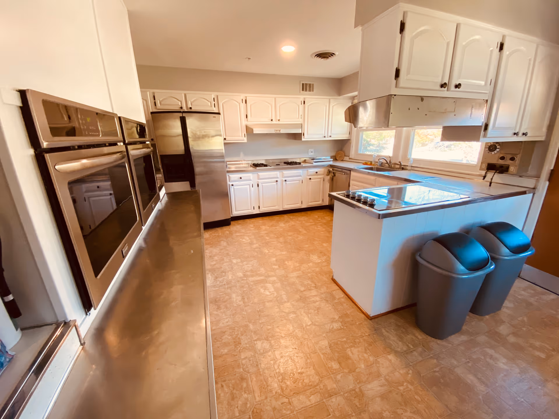 Bright commercial-style kitchen with white cabinets, stainless appliances, island, and two gray trash bins.
