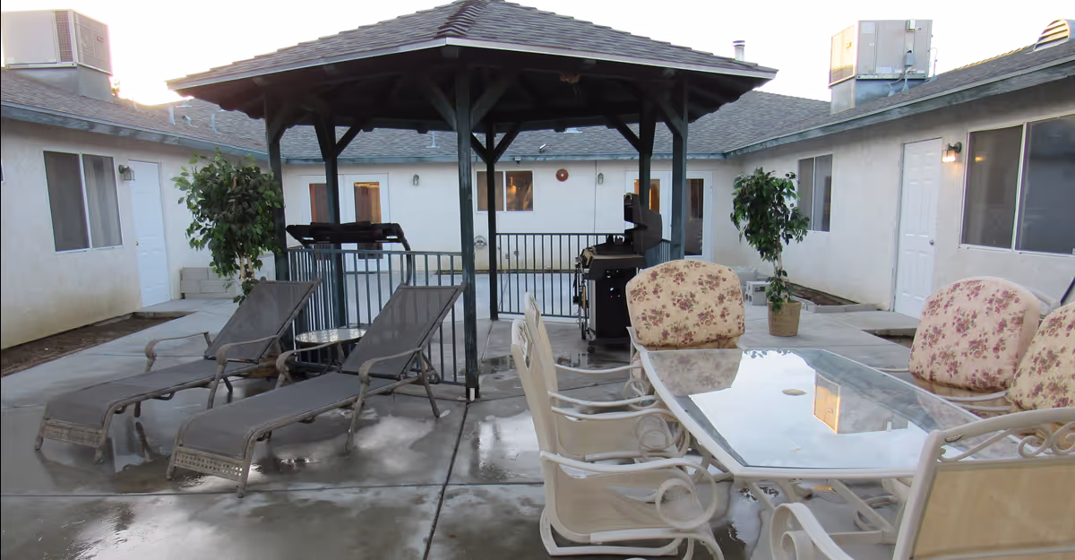 Outdoor courtyard area with a covered gazebo in the center, surrounded by a concrete patio. There are two reclining lounge chairs with a small round table between them on the left side, and a glass-top dining table with six cushioned chairs on the right side. Two potted plants are placed near the walls of the surrounding building, which has multiple doors and windows.