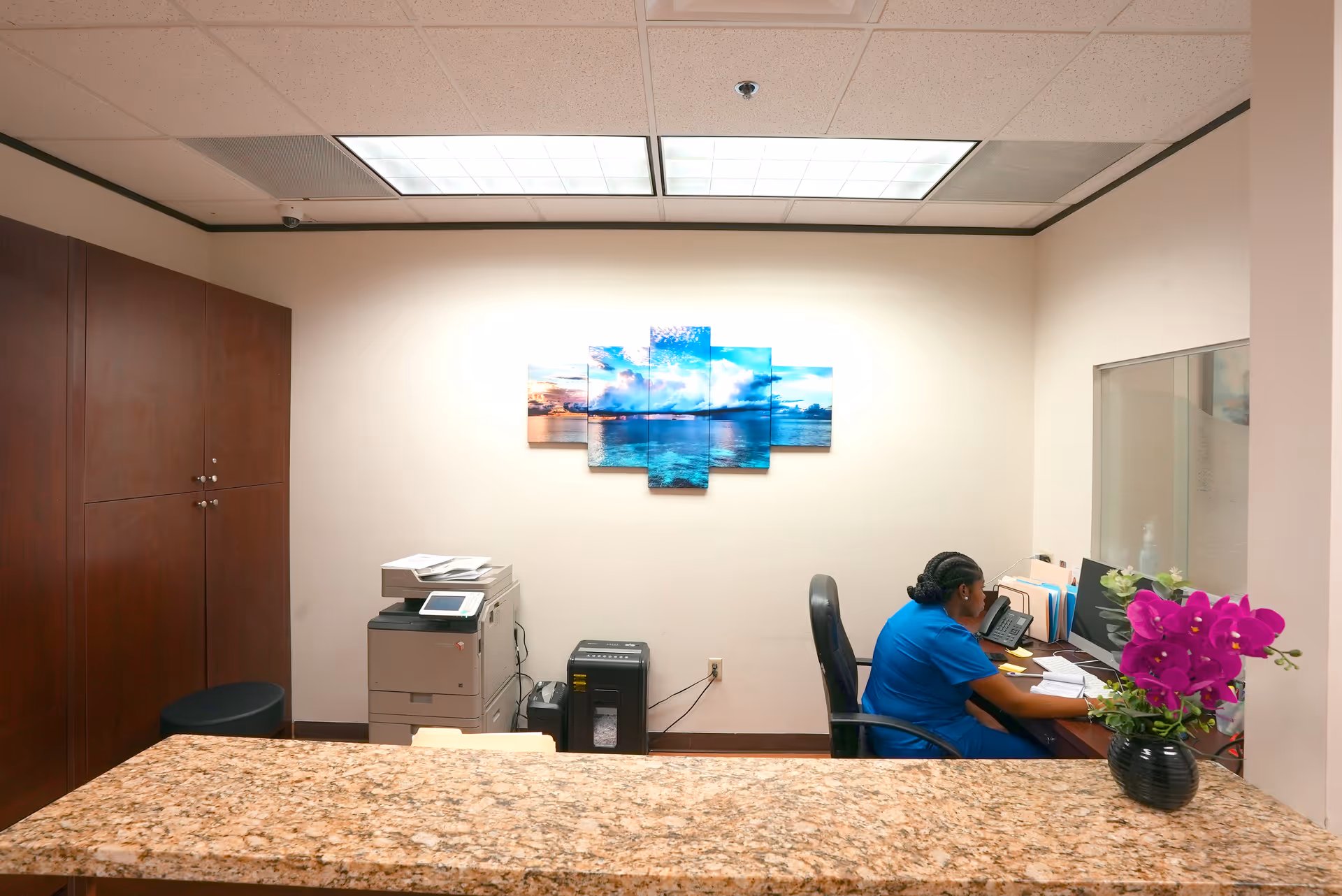 Reception area with staff at desk using computer beside a purple plant.