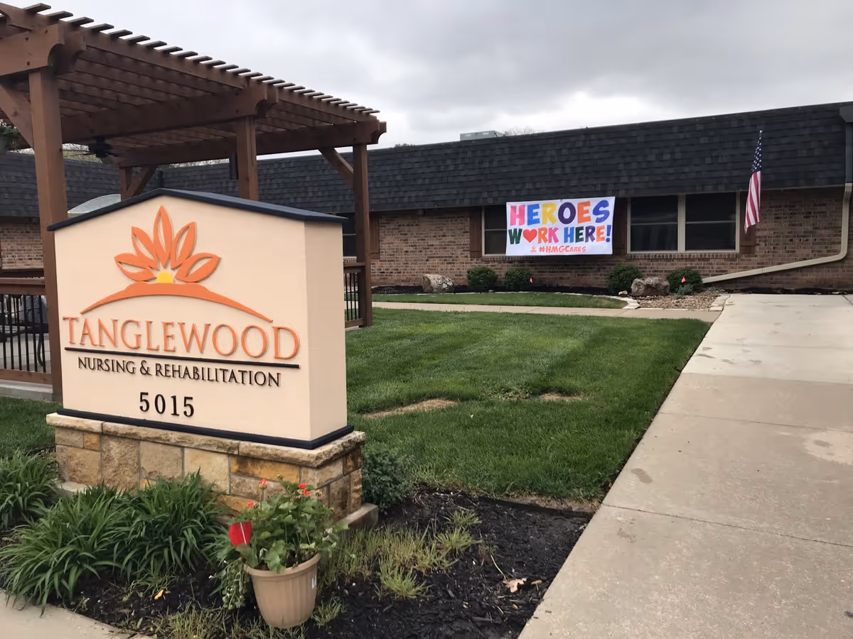 Sign reading TANGLEWOOD Nursing & Rehabilitation 5015 at the front of the facility with a lawn and a 'Heroes Work Here!' banner on the building.