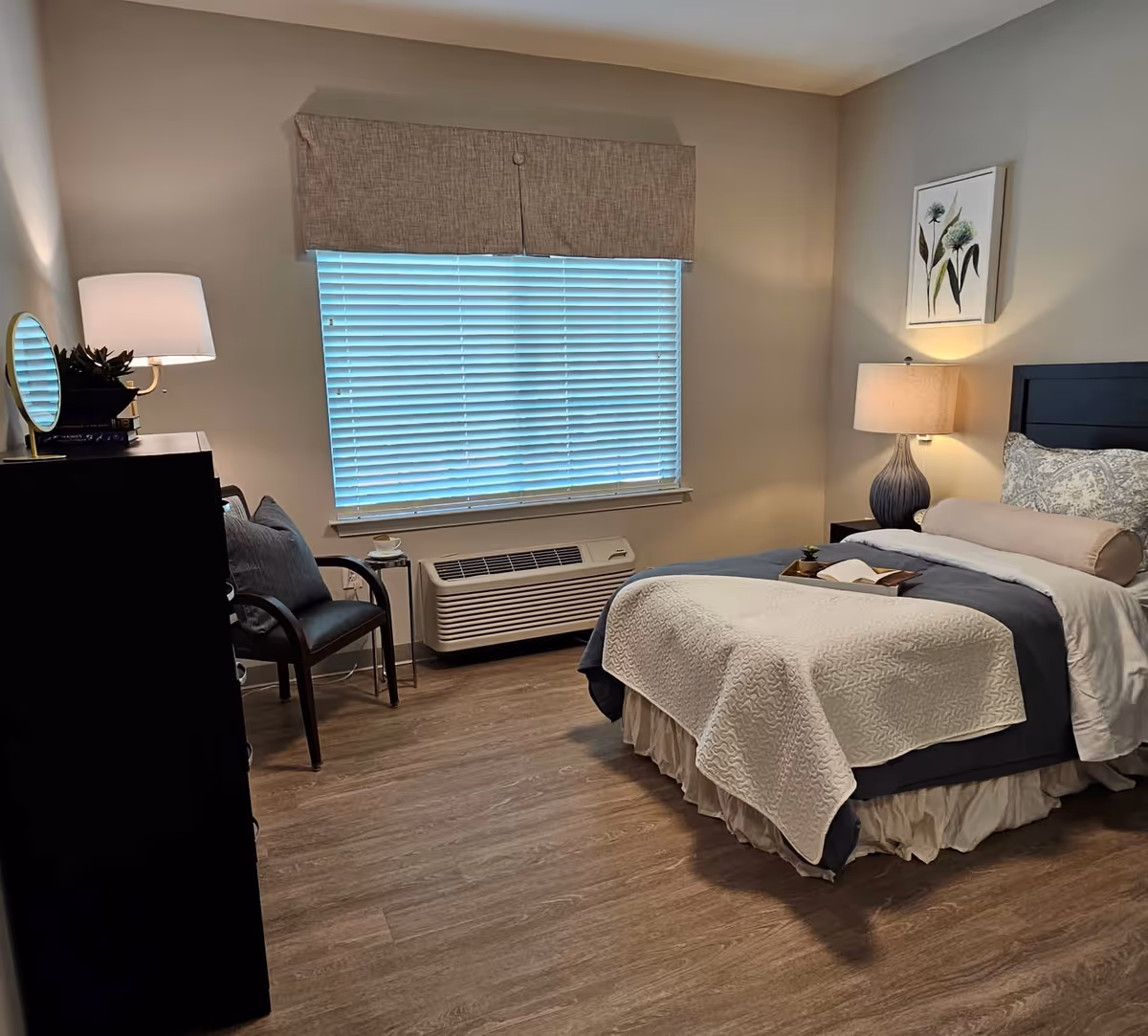 Bright hotel-style bedroom with a bed, dresser, chair, lamp, and window blinds.
