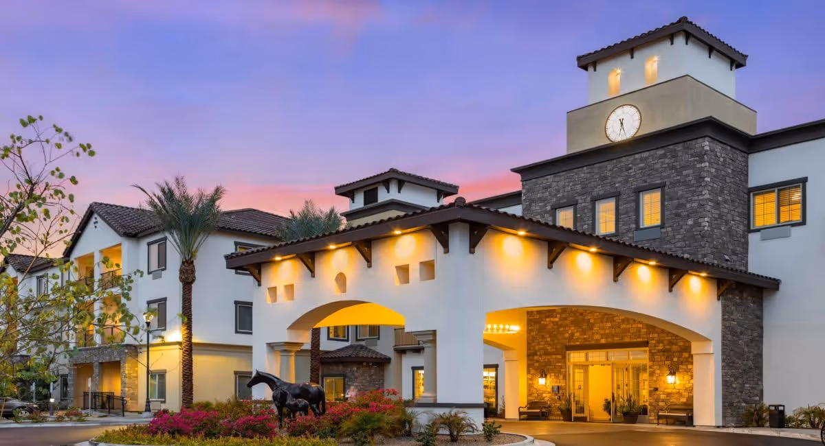 Front entrance of a Mediterranean-style senior living building at dusk with a lit canopy, clock tower, palm trees, and a horse sculpture.