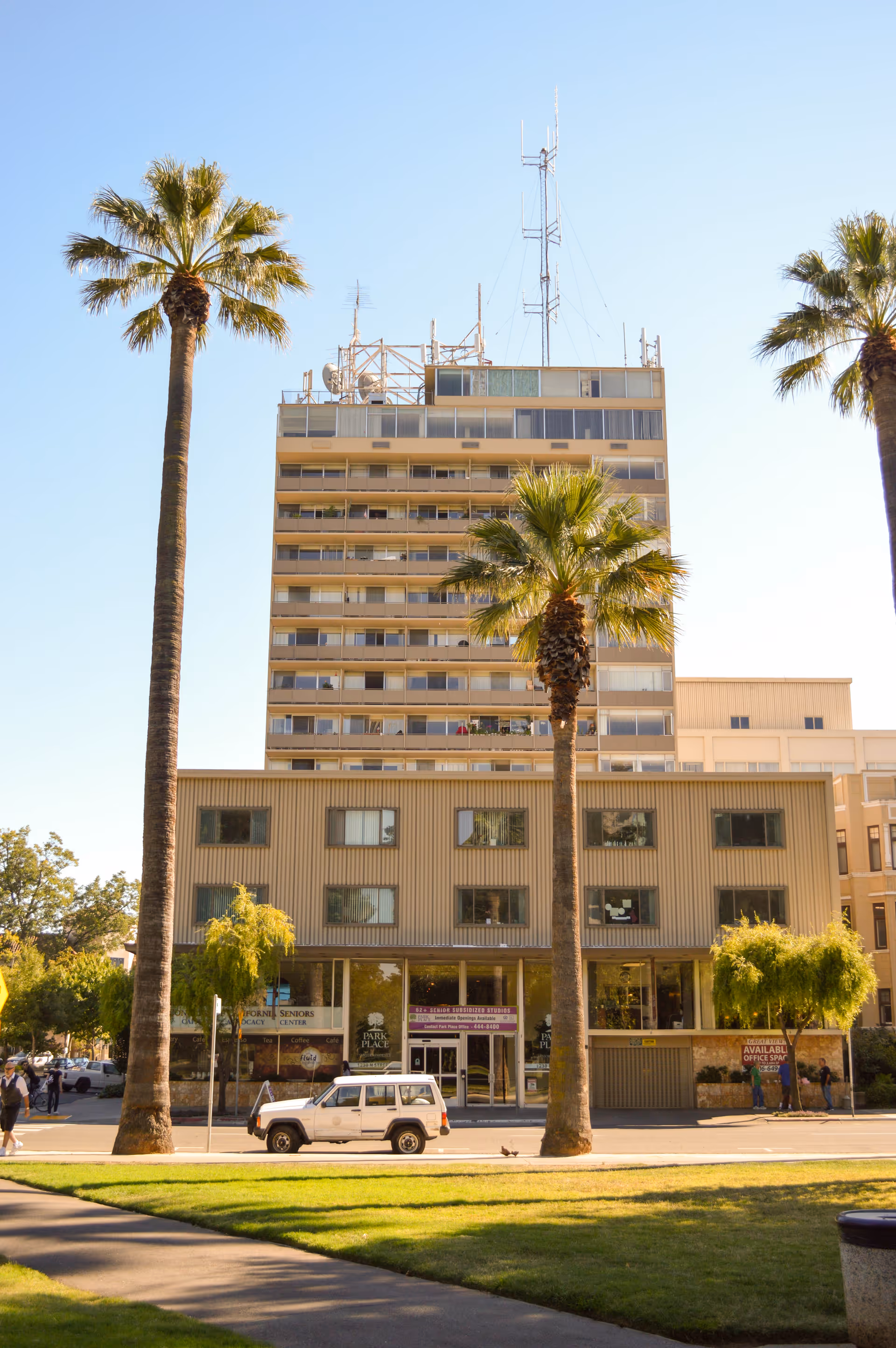 Multi-story apartment building with palm trees in front and a white SUV parked on the street.