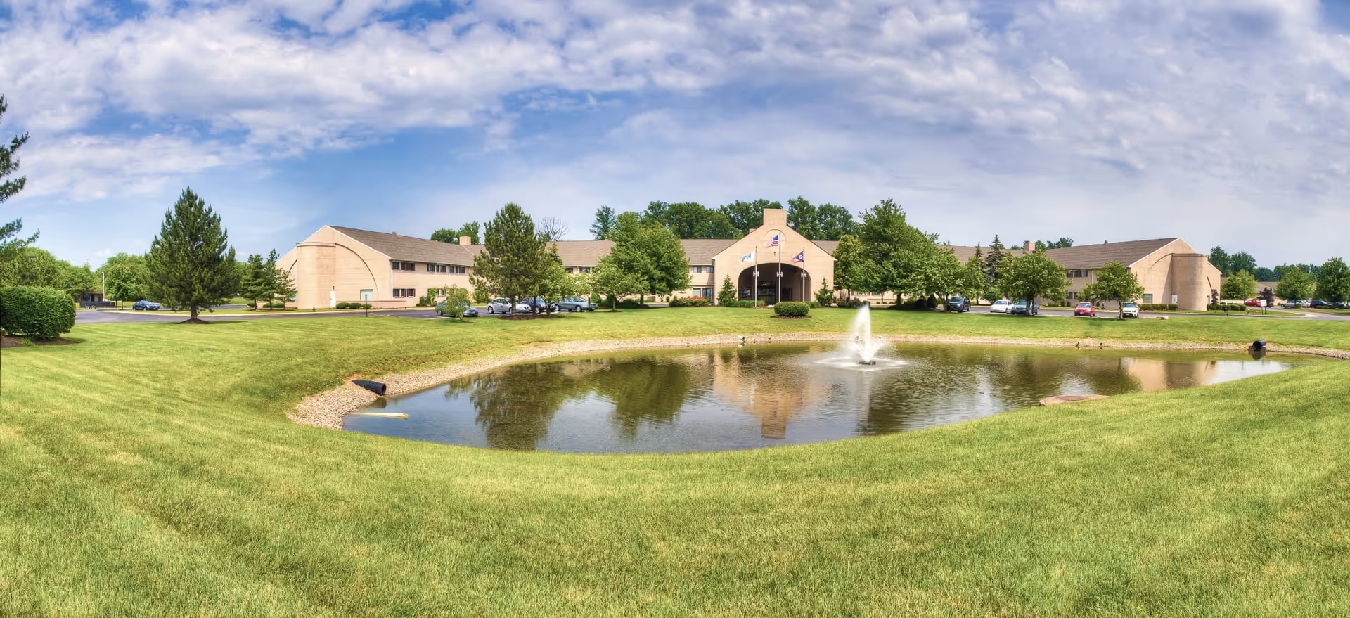 Exterior view of The Abbewood building with a central fountain pond, surrounding lawn and trees under a partly cloudy sky.