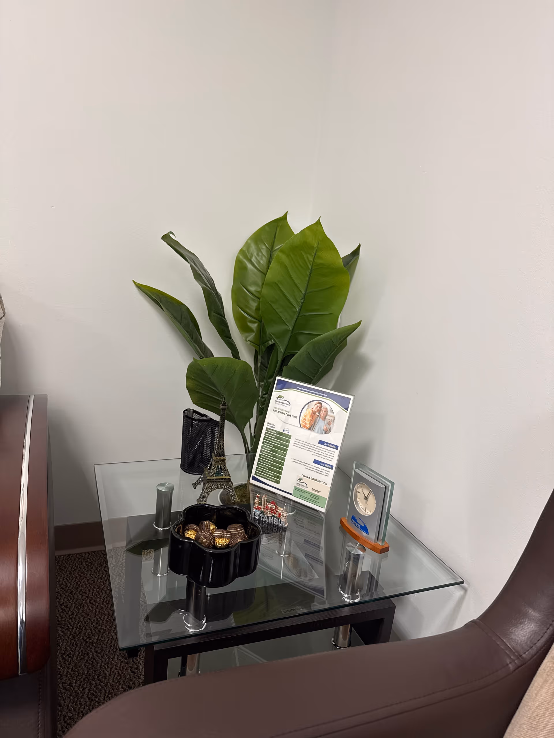 Glass coffee table with chocolates, plant, brochure, clock, and chair in a lobby area.