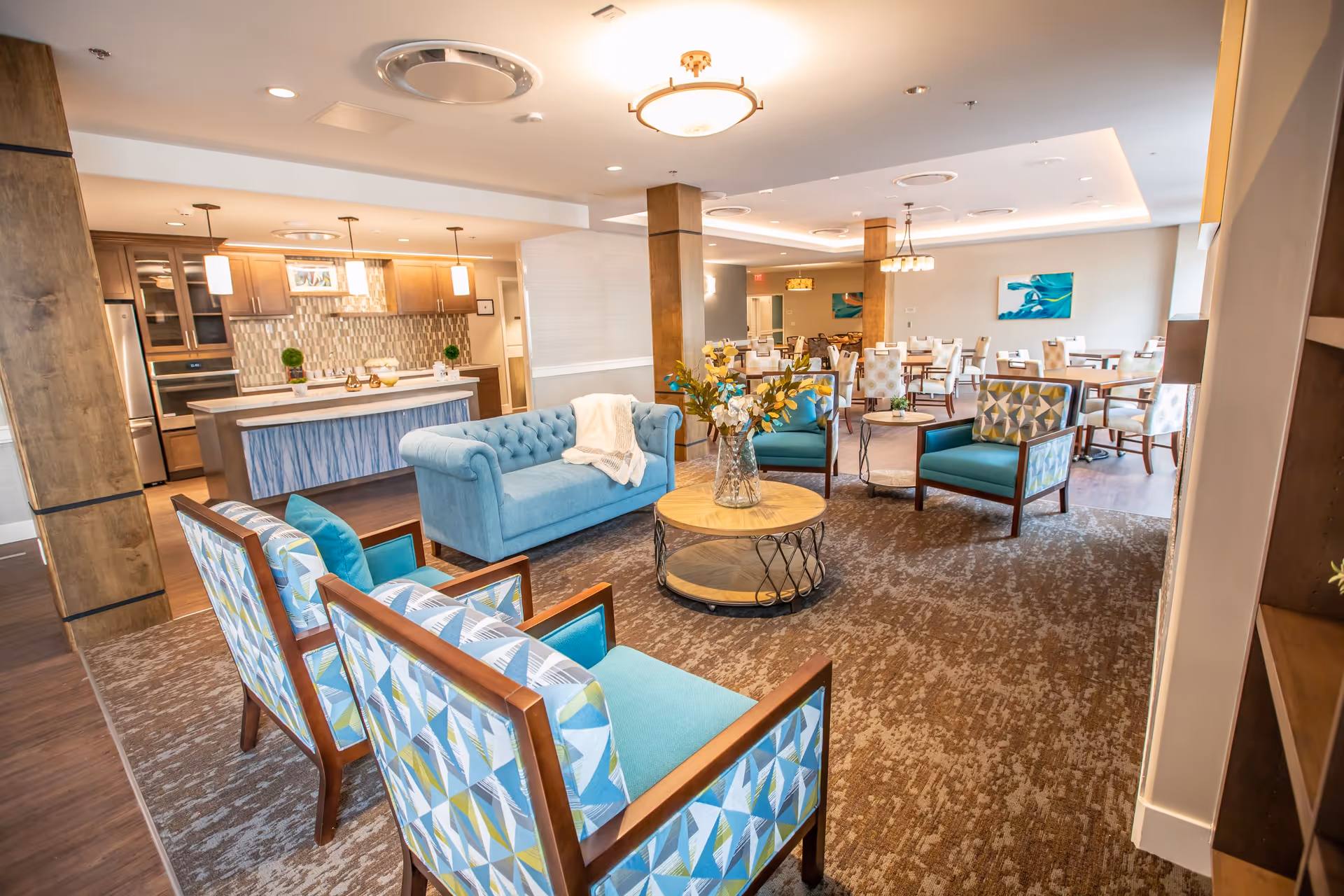 Bright senior living lounge with teal chairs, a blue sofa, and a kitchen in background