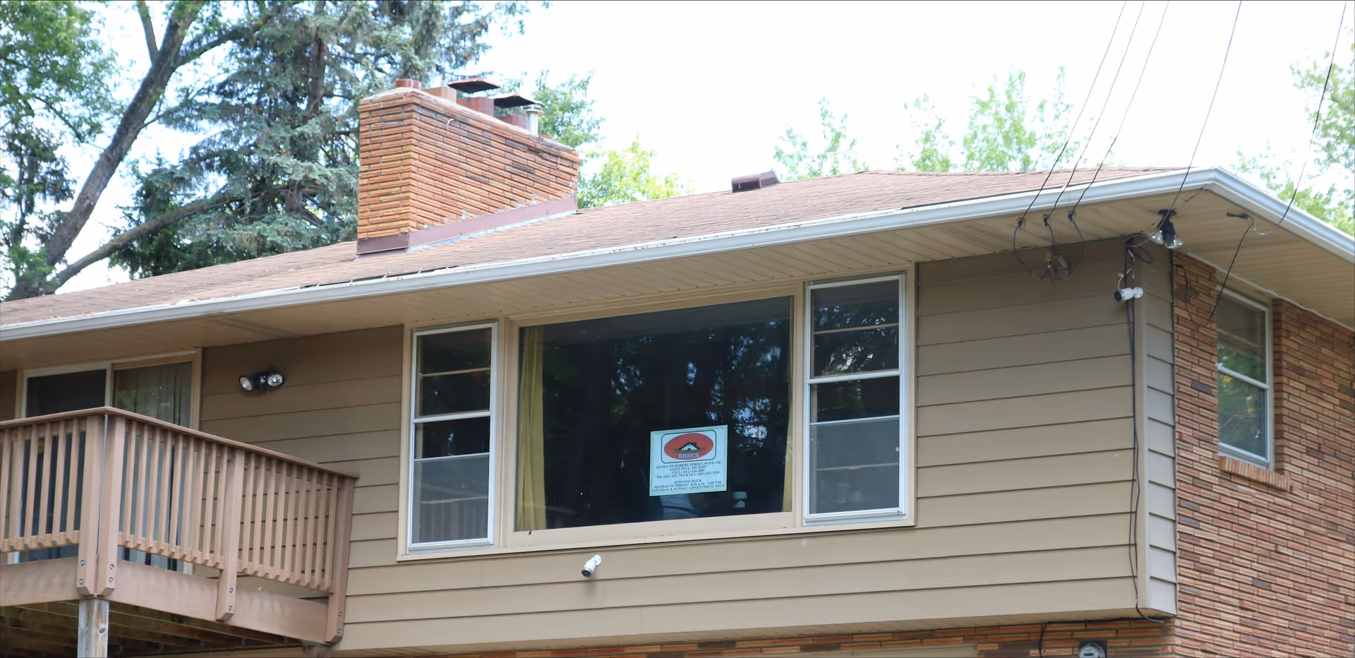 Two-story beige house with a wooden balcony, chimney, and large front windows.