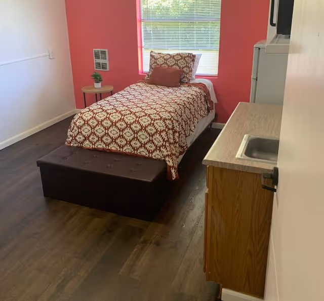 A small bedroom with a patterned bedspread, burgundy wall, window, and kitchenette counter.