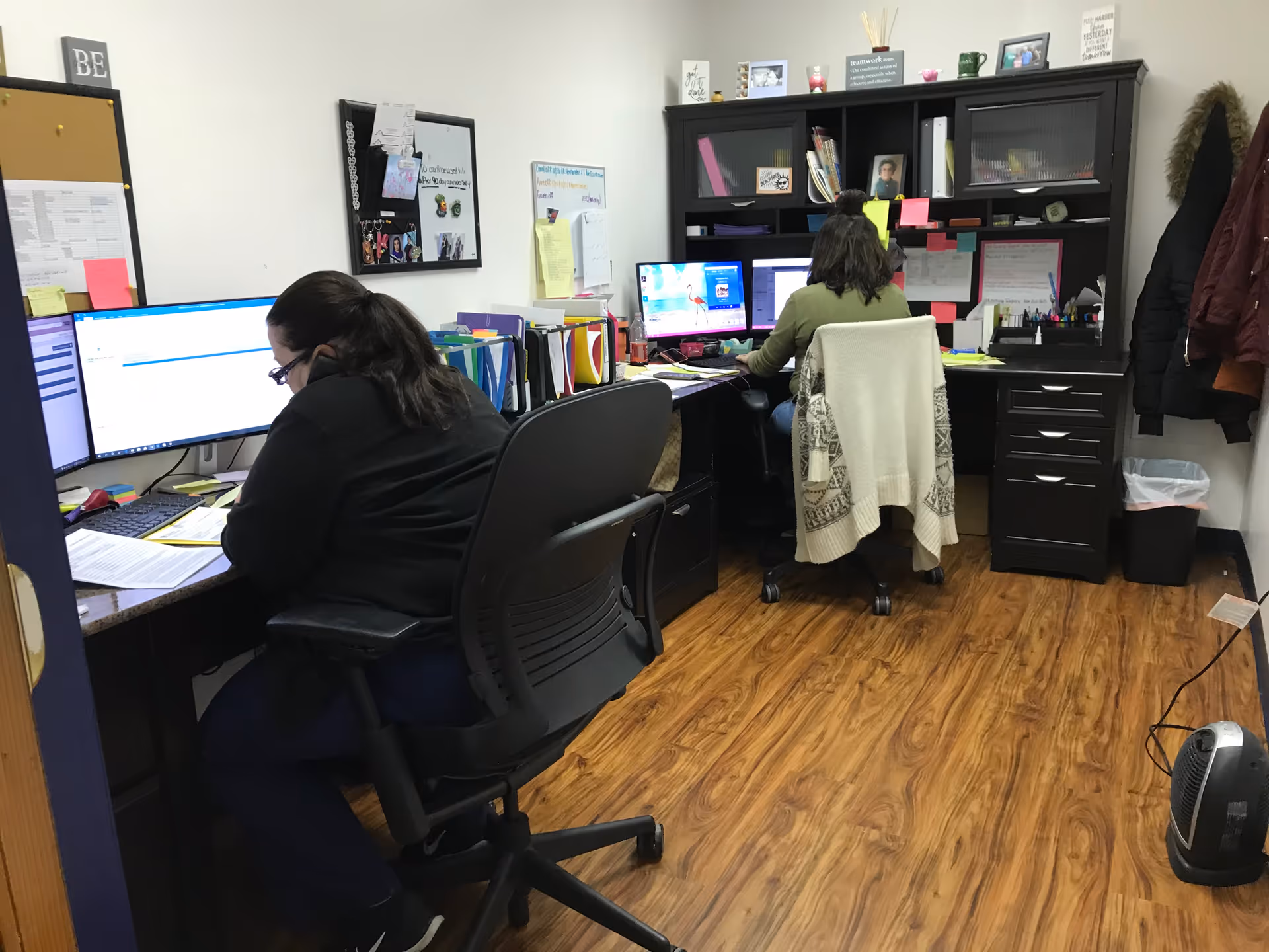 Two office workers sit at desks with computers and organized supplies in a home care office.