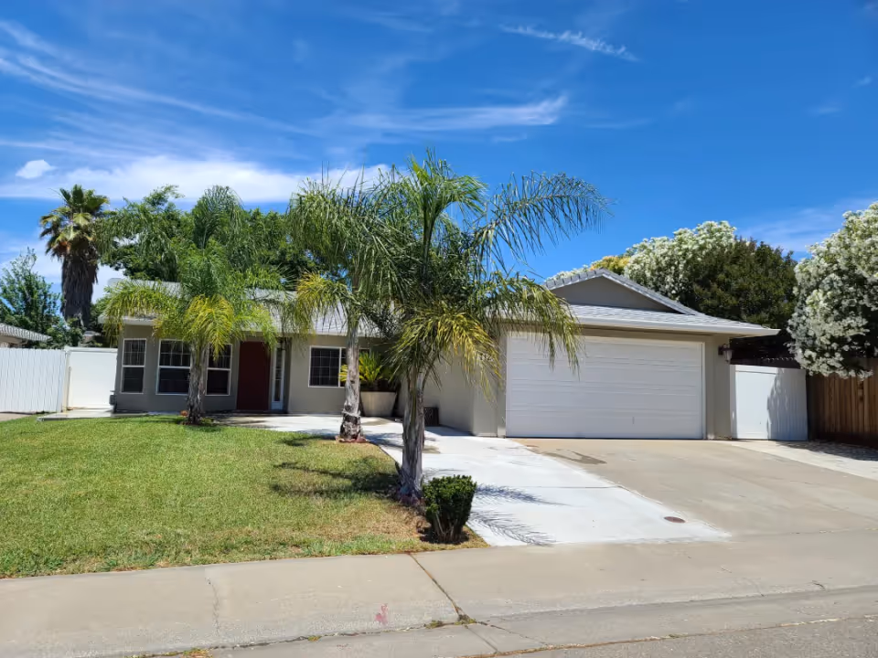 Front view of a single-story suburban house with a two-car garage, driveway, lawn and palm trees under a blue sky.