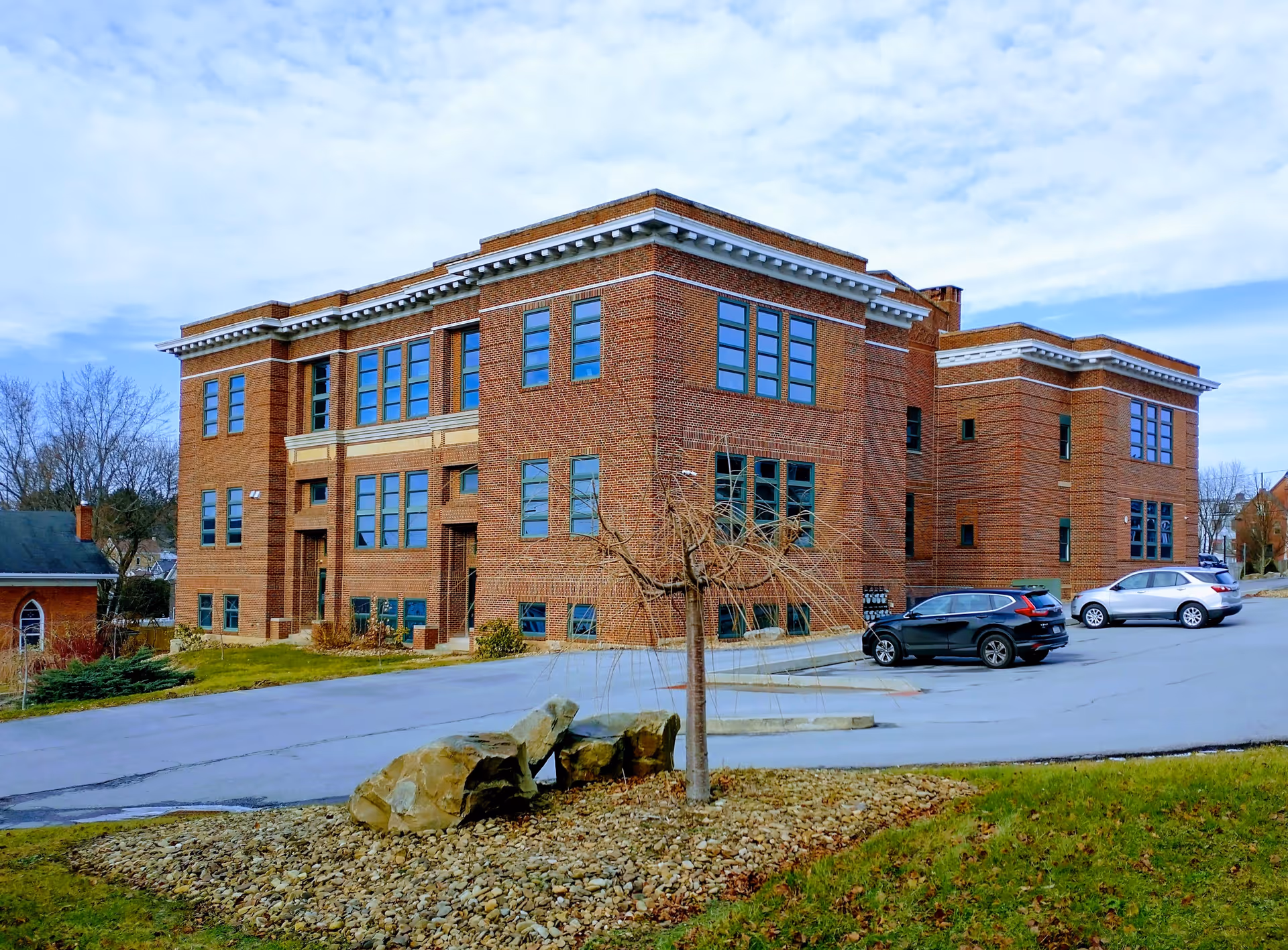 Brick multi-story senior living facility exterior with cars in a driveway and a bare tree