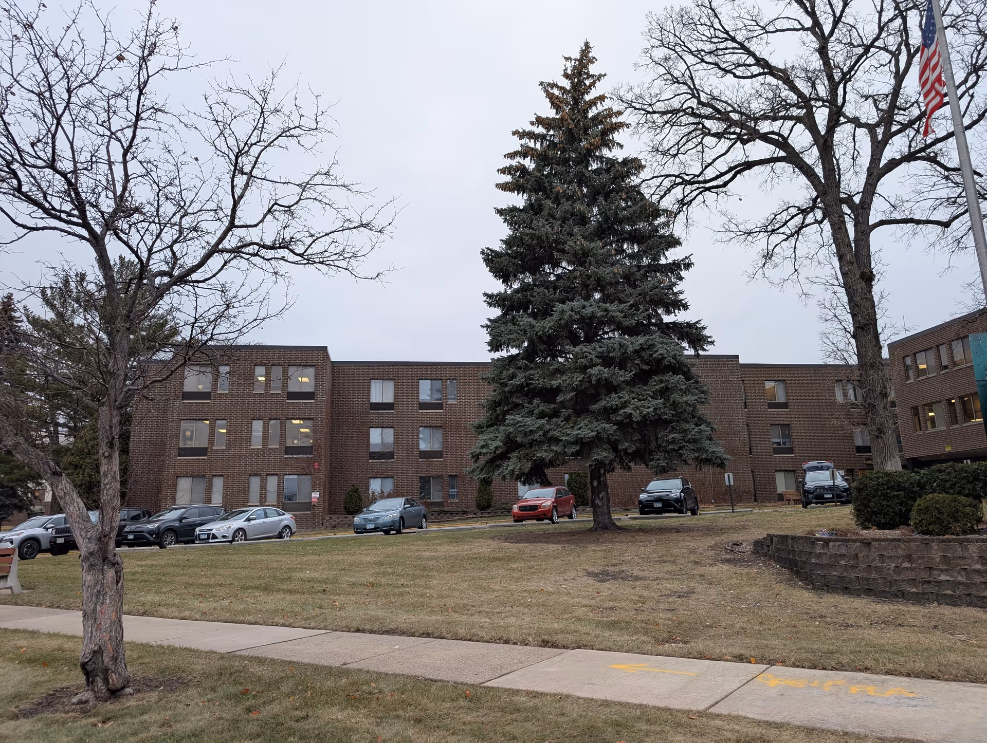 Exterior view of a three-story brick building with several windows, surrounded by a grassy area with a large evergreen tree and leafless deciduous trees. Several cars are parked in front of the building, and an American flag is visible on a flagpole to the right.