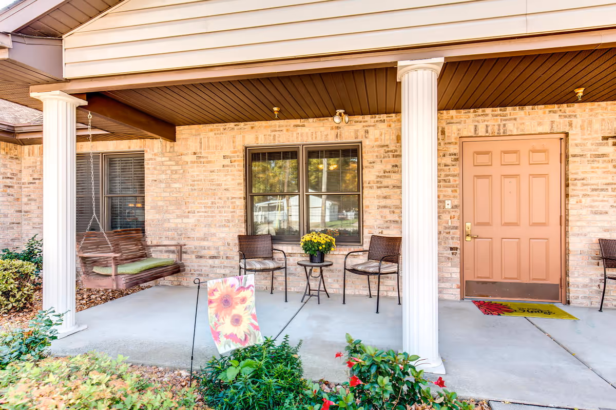 Covered porch area with two white columns, a wooden swing with green cushions hanging from the ceiling, two brown chairs with cushions, a small round table with a yellow flower pot, a window with blinds, a brown door with a welcome mat, and some green plants and flowers in the foreground.
