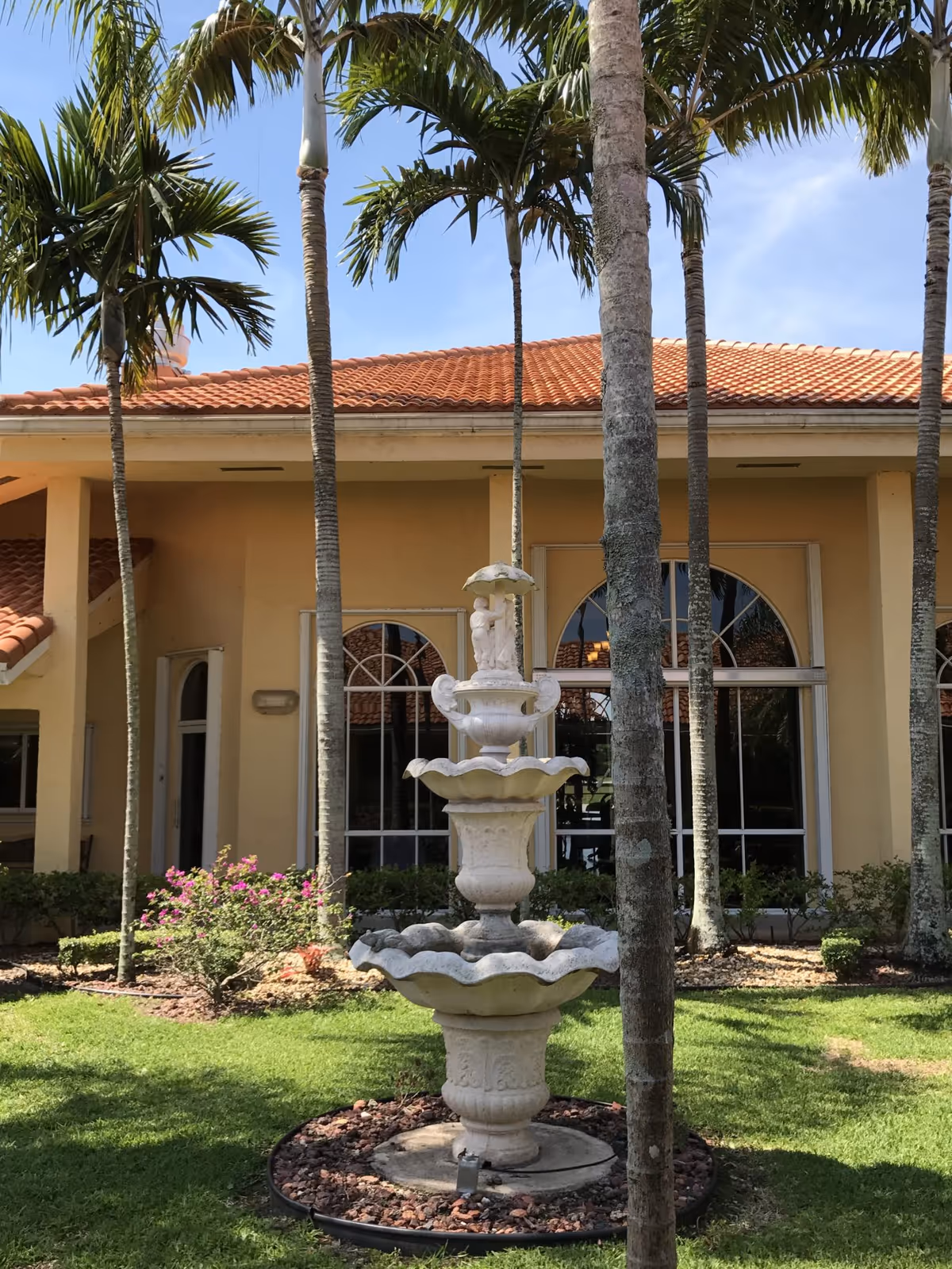 Tiered stone fountain in a grassy, palm‑lined courtyard in front of a Mediterranean-style building with arched windows.