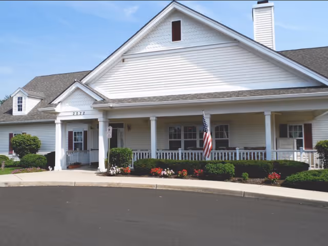 White senior living facility front with porch, shrubs, and American flag.