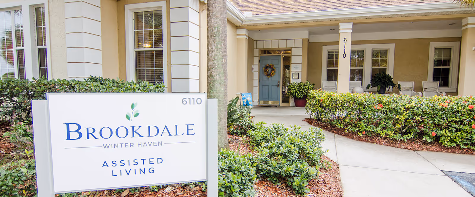 Exterior view of the entrance to Brookdale Winter Haven Assisted Living facility, showing a sign with the facility name and address number 6110, surrounded by greenery and a walkway leading to a covered porch with chairs and plants.