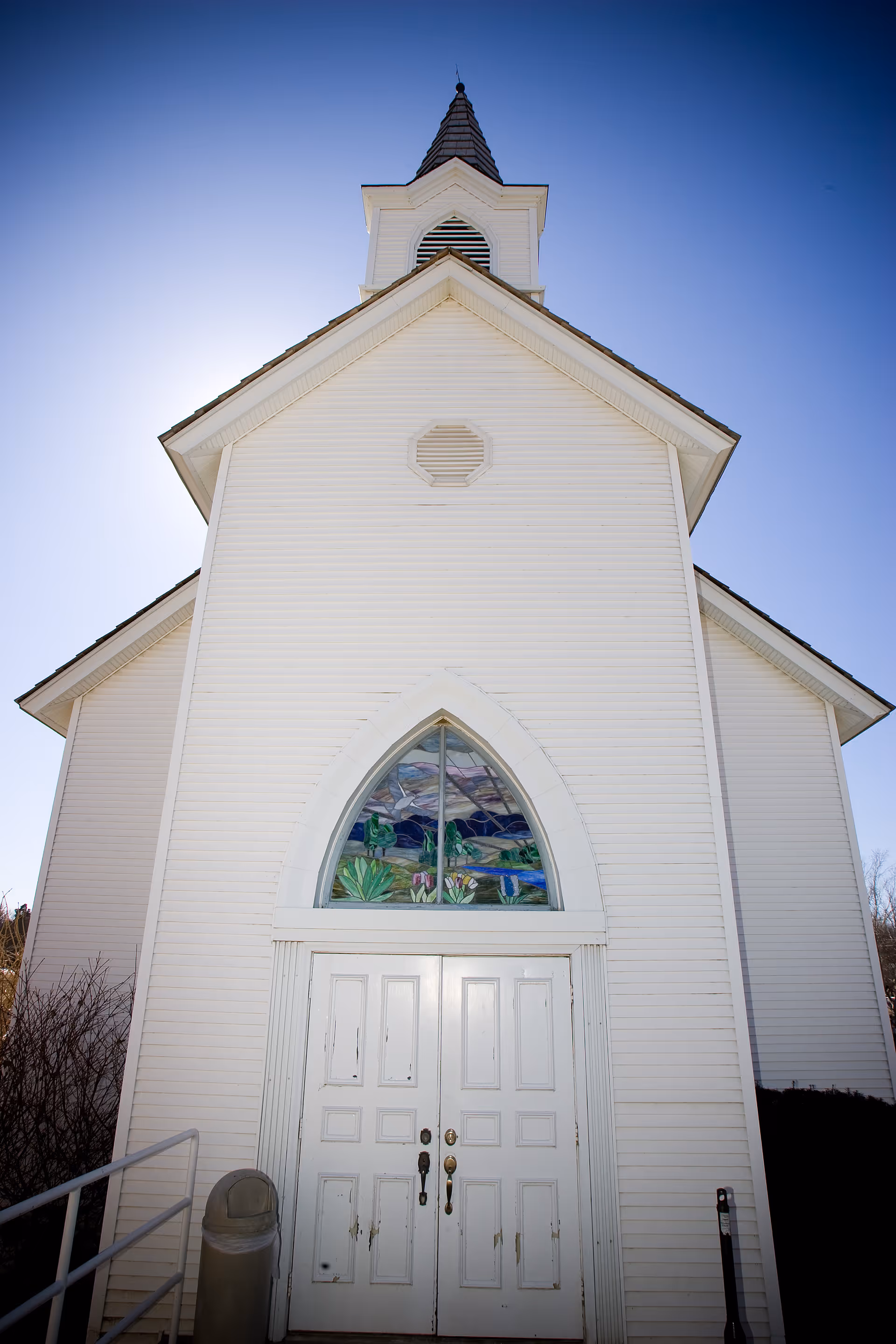 Front exterior of a white church-like building with double doors, an arched stained-glass window above them, and a steeple against a clear blue sky.