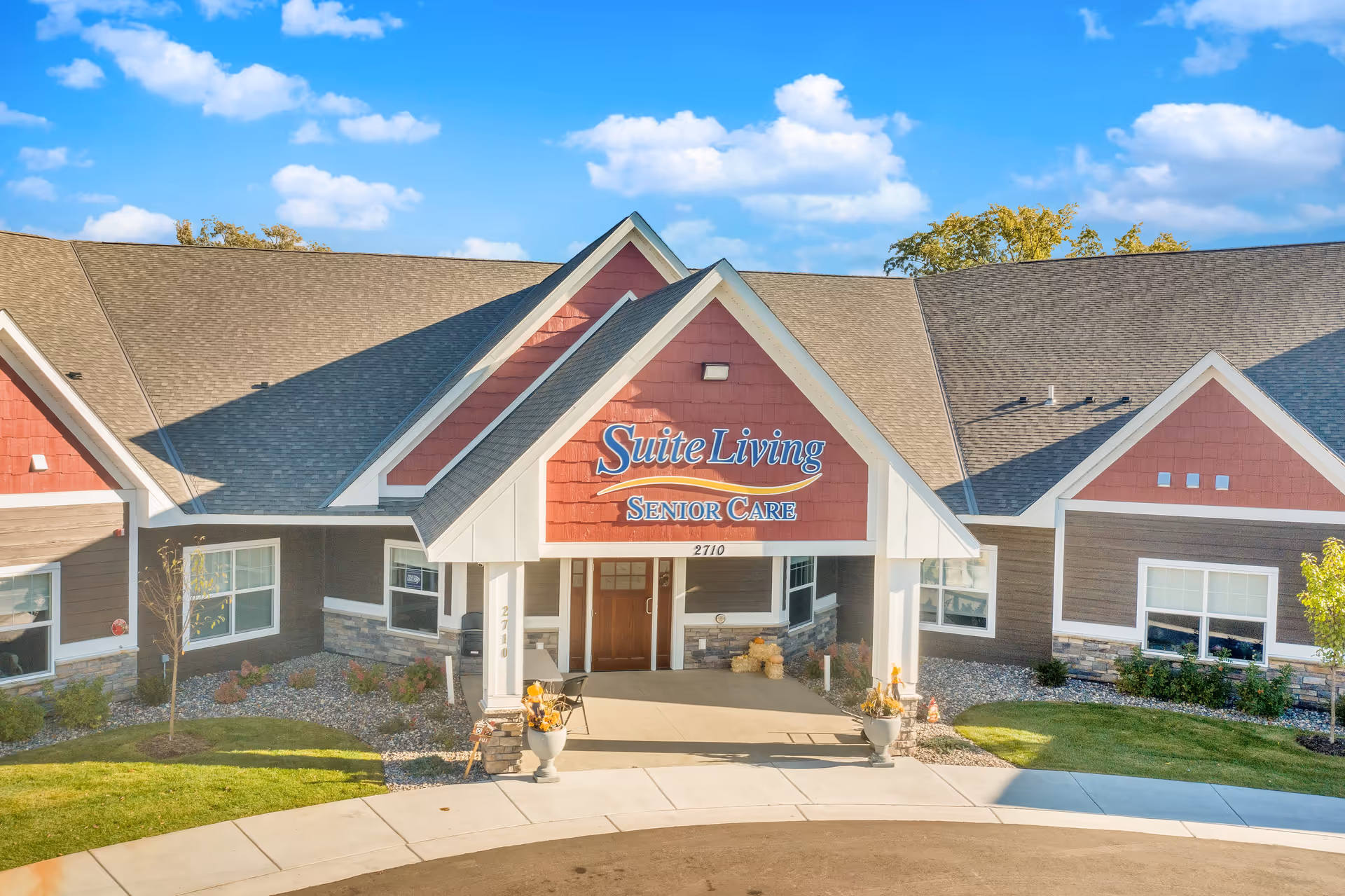 Front exterior view of Suite Living Senior Care facility with a peaked roof, red and brown siding, stone accents, and a covered entrance. The building is surrounded by a landscaped area with grass, small trees, and shrubs under a blue sky with scattered clouds.
