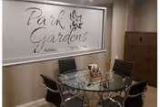 A glass round table with four chairs sits in a wood-paneled interior beside Park Gardens sign.