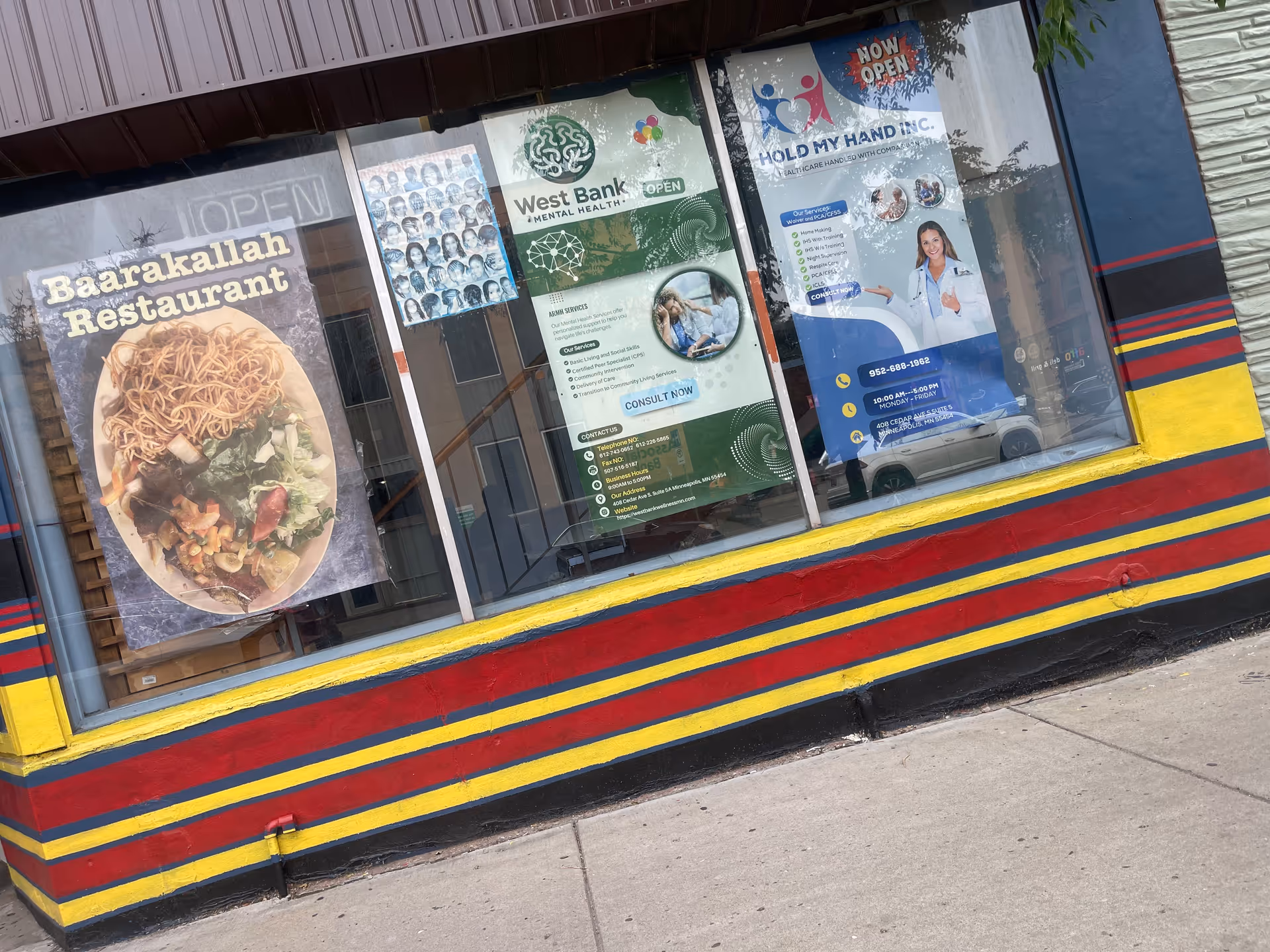 Storefront with colorful posters in large windows advertising businesses, including Hold My Hand Inc.