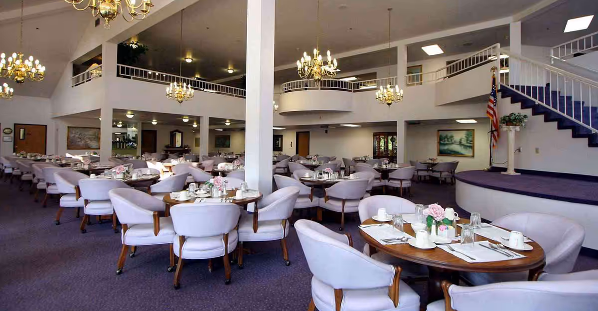 Large dining room with round tables set for meals, upholstered chairs, chandeliers, and a raised stage with stairs.