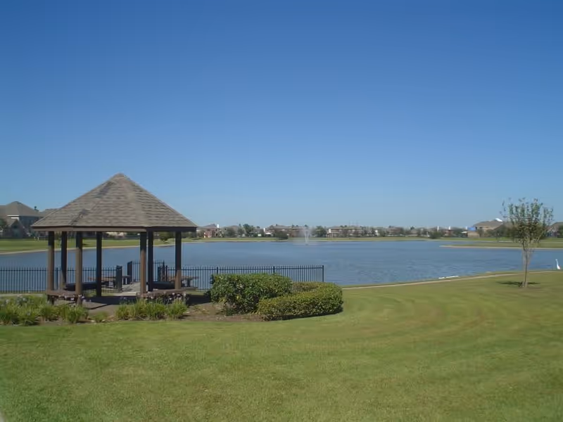 A peaceful outdoor scene featuring a wooden gazebo with a shingled roof near a fenced pond with a water fountain in the distance. The area is surrounded by well-maintained green grass, bushes, and a few small trees under a clear blue sky.