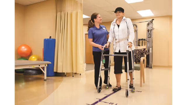 A physical therapist assists an elderly woman using a walker in a rehabilitation room with exercise equipment and therapy balls in the background.