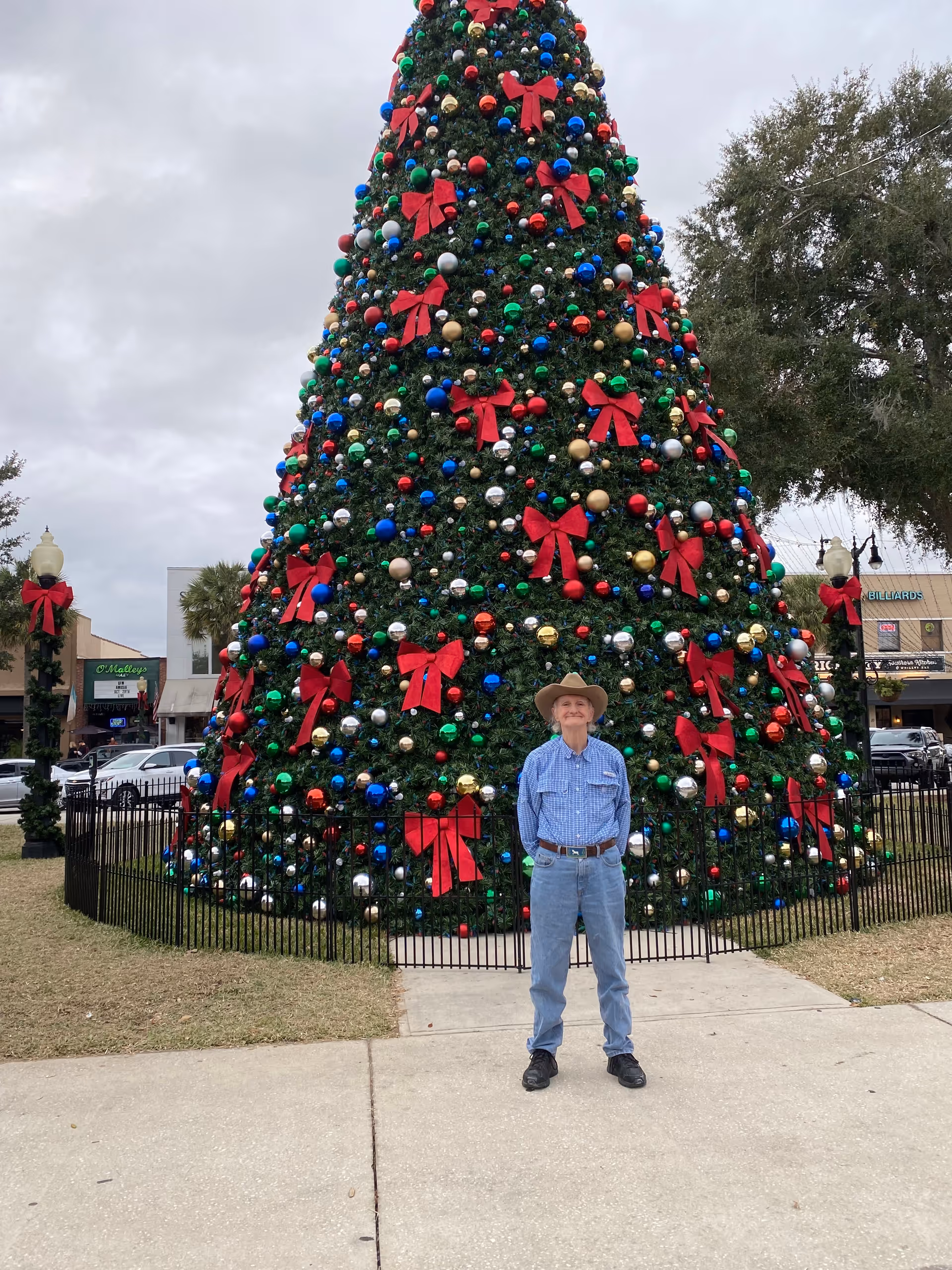 A man in a cowboy hat stands in front of a large decorated Christmas tree outdoors.