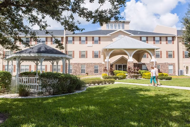 Front exterior of a multistory retirement community with a covered entrance, a white gazebo on the lawn, and two people walking on a pathway.