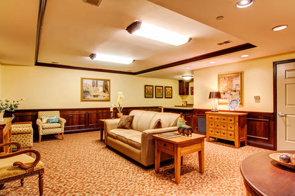 A cozy living room area with a beige sofa, wooden side tables, and several armchairs. The room features warm lighting from ceiling fixtures and a table lamp, framed artwork on the walls, and a patterned carpet. There is a wooden cabinet and a door on the right side of the room.