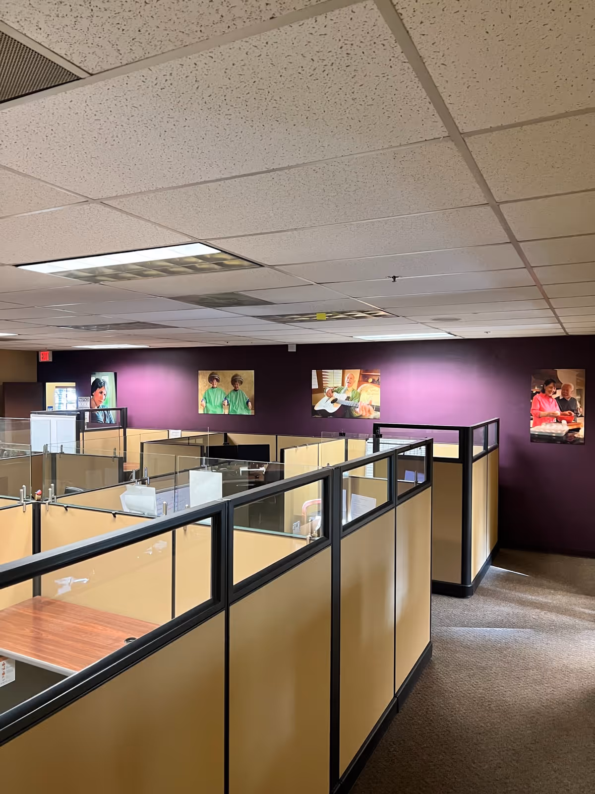 Interior view of a cubicle office area with beige partitions and purple walls featuring posters.