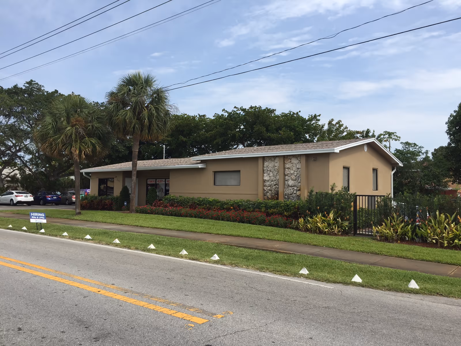 Single-story beige facility building with manicured shrubs, palm trees, and a sidewalk along a street.