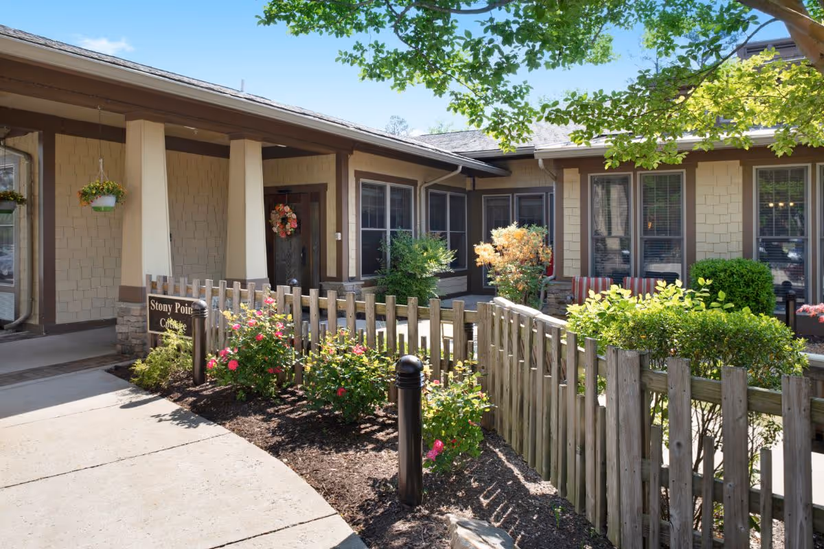 Exterior view of a single-story building with beige siding and brown trim, featuring a small fenced garden area with blooming flowers and shrubs. A pathway leads to a door decorated with a floral wreath. Trees with green leaves provide shade.