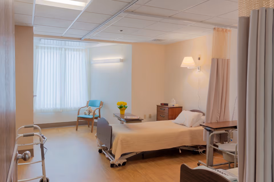 Hospital-style patient room with a bed, privacy curtains, chair, and bedside table.
