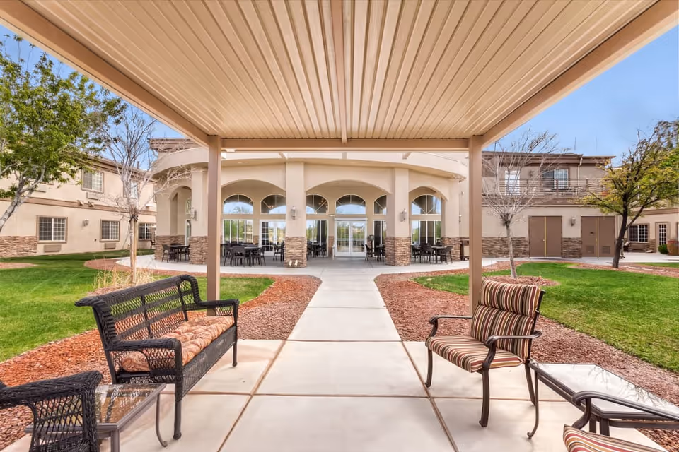Covered patio with chairs and benches overlooking a landscaped courtyard and the front entrance of a two-story senior living building.