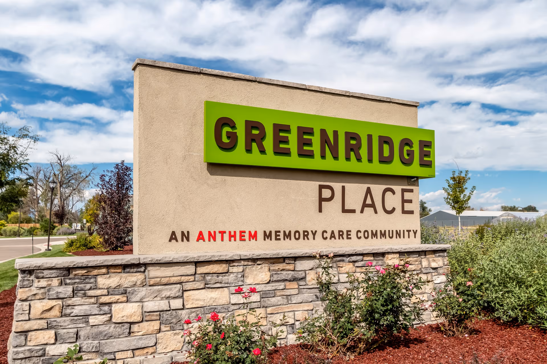 Outdoor sign for Greenridge Place, an Anthem memory care community, with a stone base and landscaping including red flowers and bushes under a partly cloudy sky.