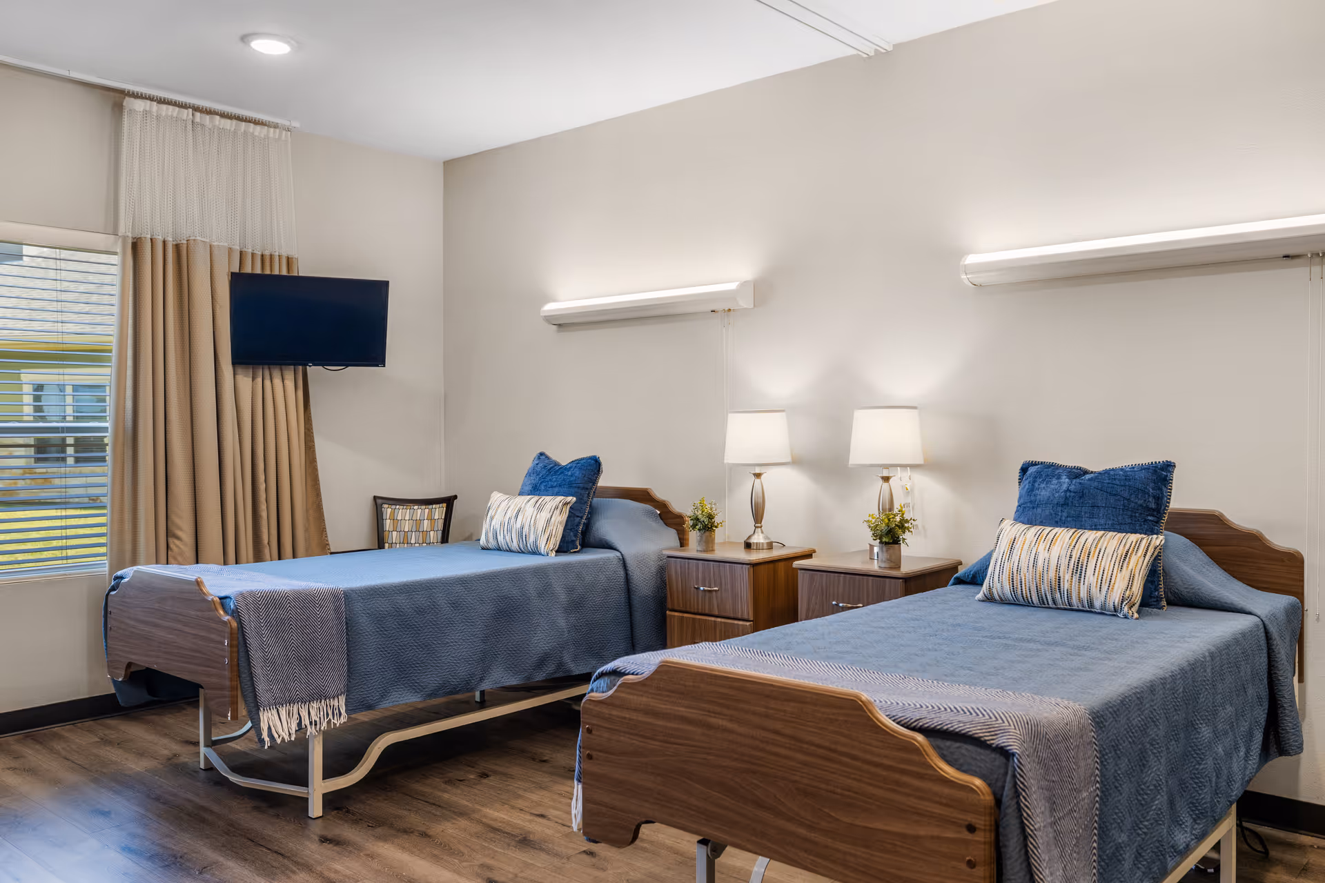 Two single hospital beds with blue bedding and nightstands in a care facility bedroom.