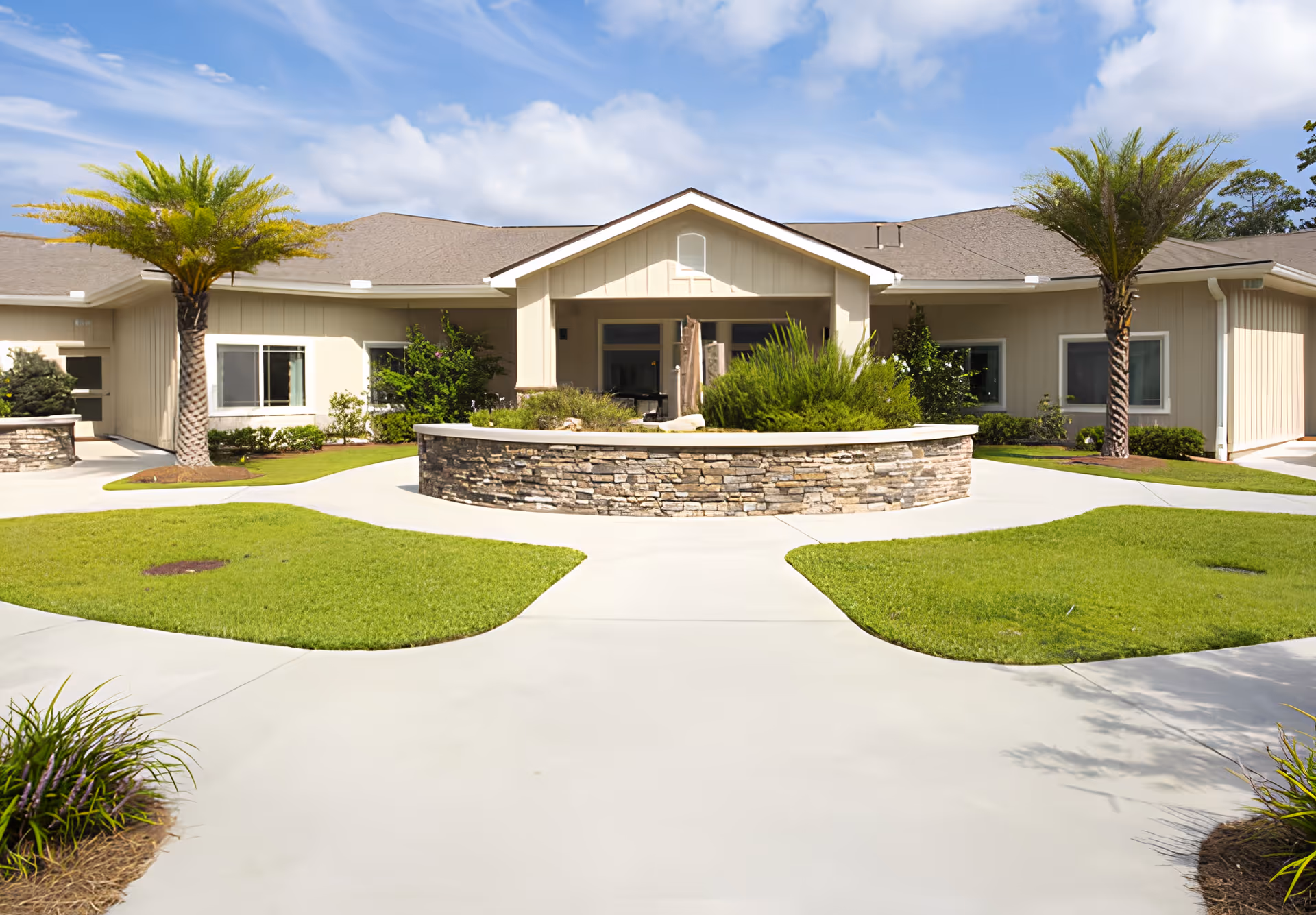Front exterior view of Azalea Gardens Assisted Living & Memory Care building with a stone planter in the center, green lawns, palm trees, and a clear blue sky with some clouds.
