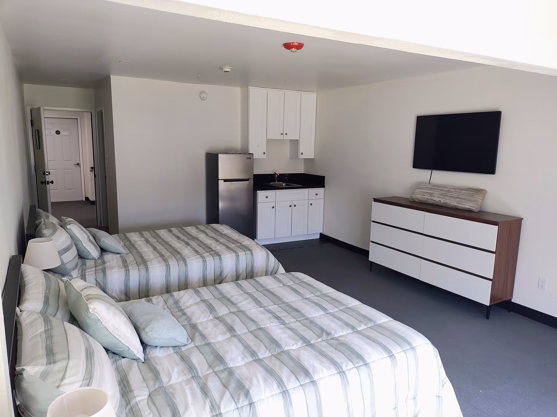 Two beds, a kitchenette, and a dresser with a mounted TV in a senior living suite.
