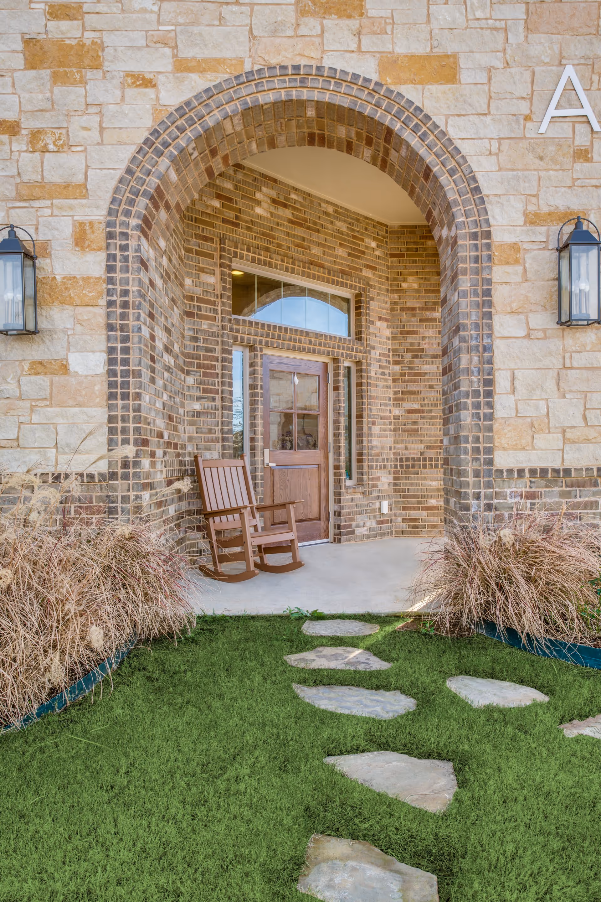 Stone and brick building entrance with an arched doorway, a wooden door with glass panels, a wooden rocking chair on the porch, two wall-mounted lantern lights, and a stone pathway leading through green grass to the entrance, flanked by ornamental grasses.
